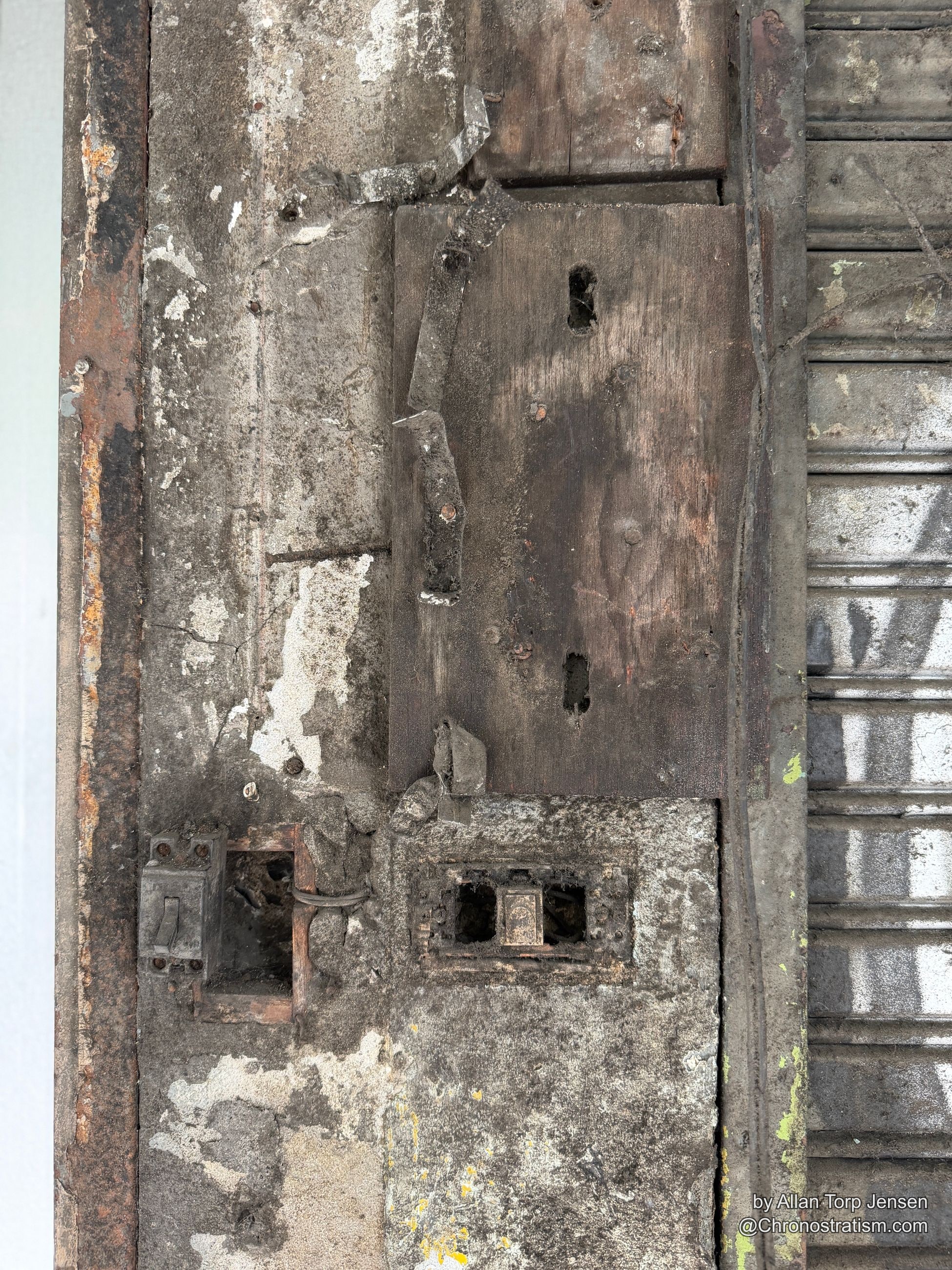 A charred, soot-darkened wood door panel with corroded electrical fixtures and a rusted latch.