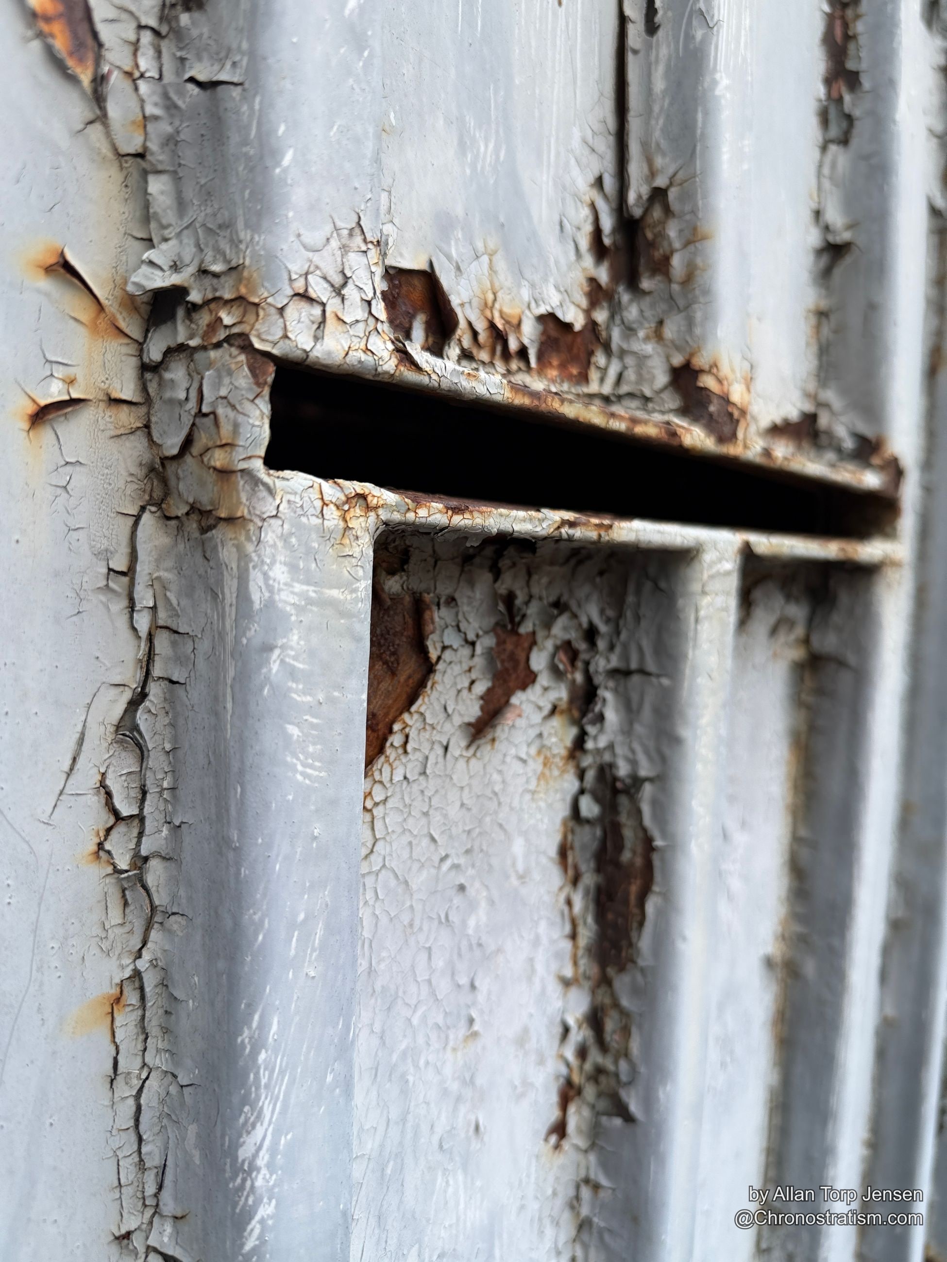 Close-up of a rusted mail slot edge, cracked white paint radiating outward.