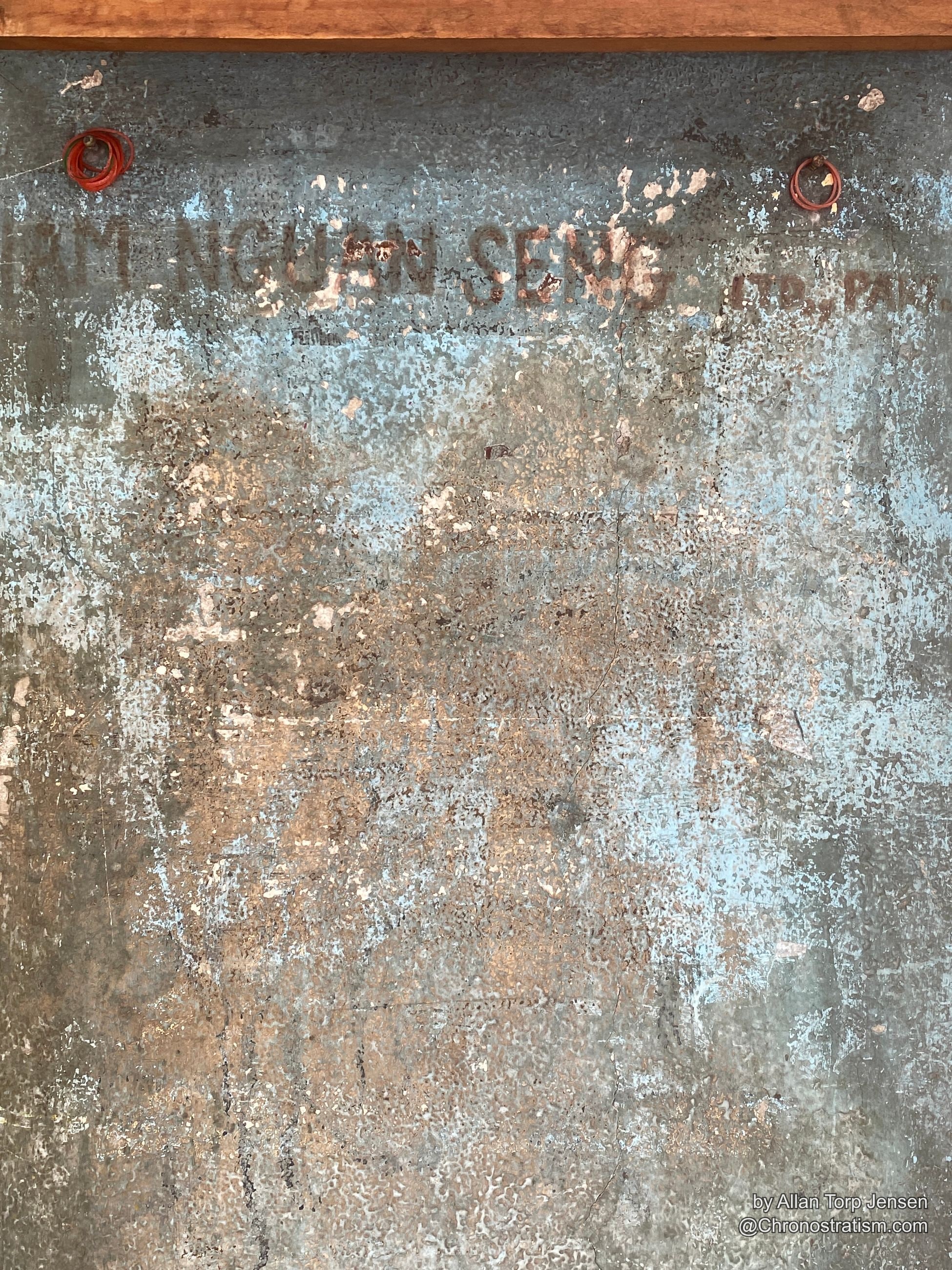 Faded ghost-sign lettering bleeds through decades of peeling paint on a weathered metal panel.