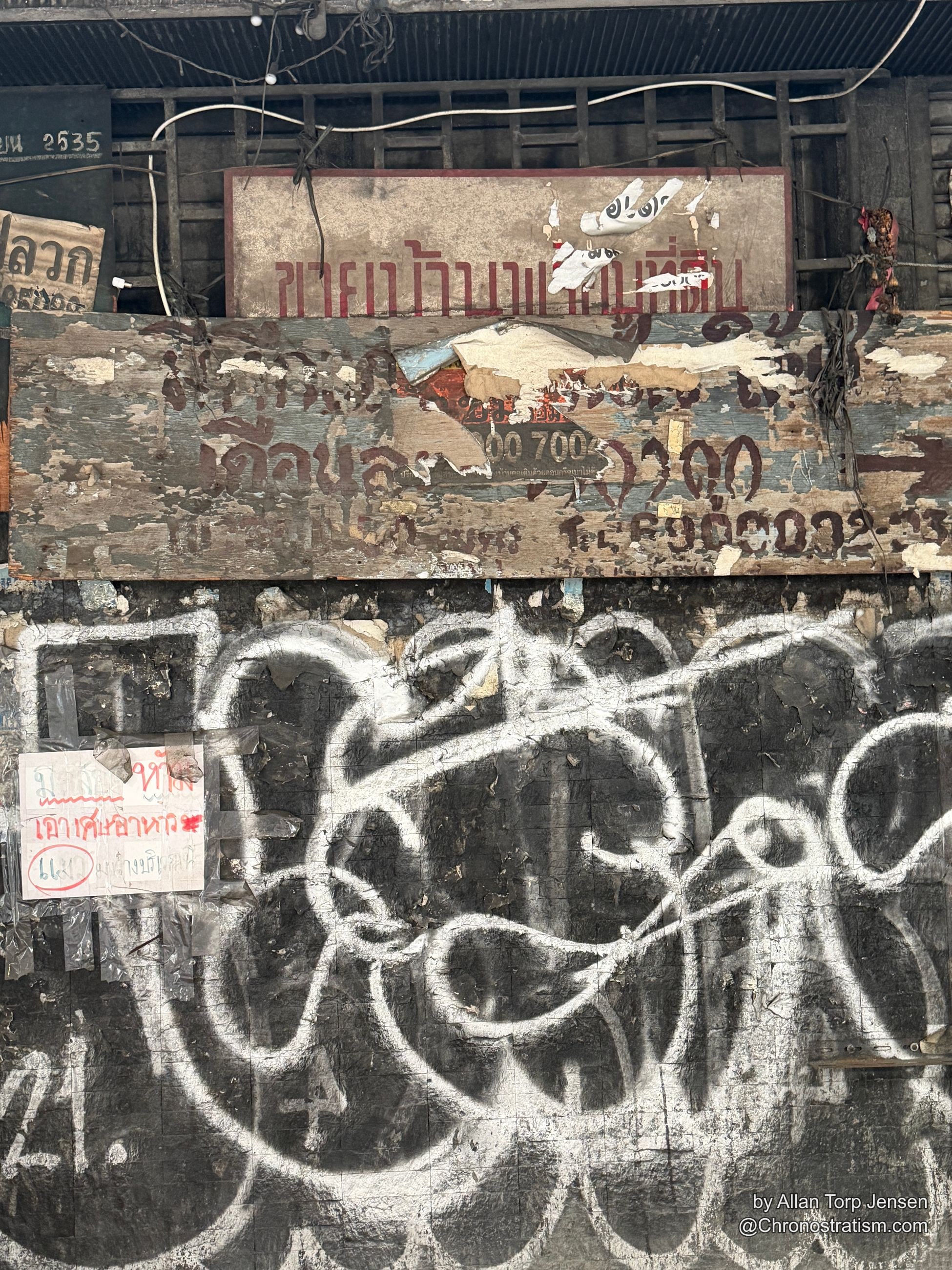 Faded maroon Thai lettering peels from an old wooden signage board beside a spray-painted mural.