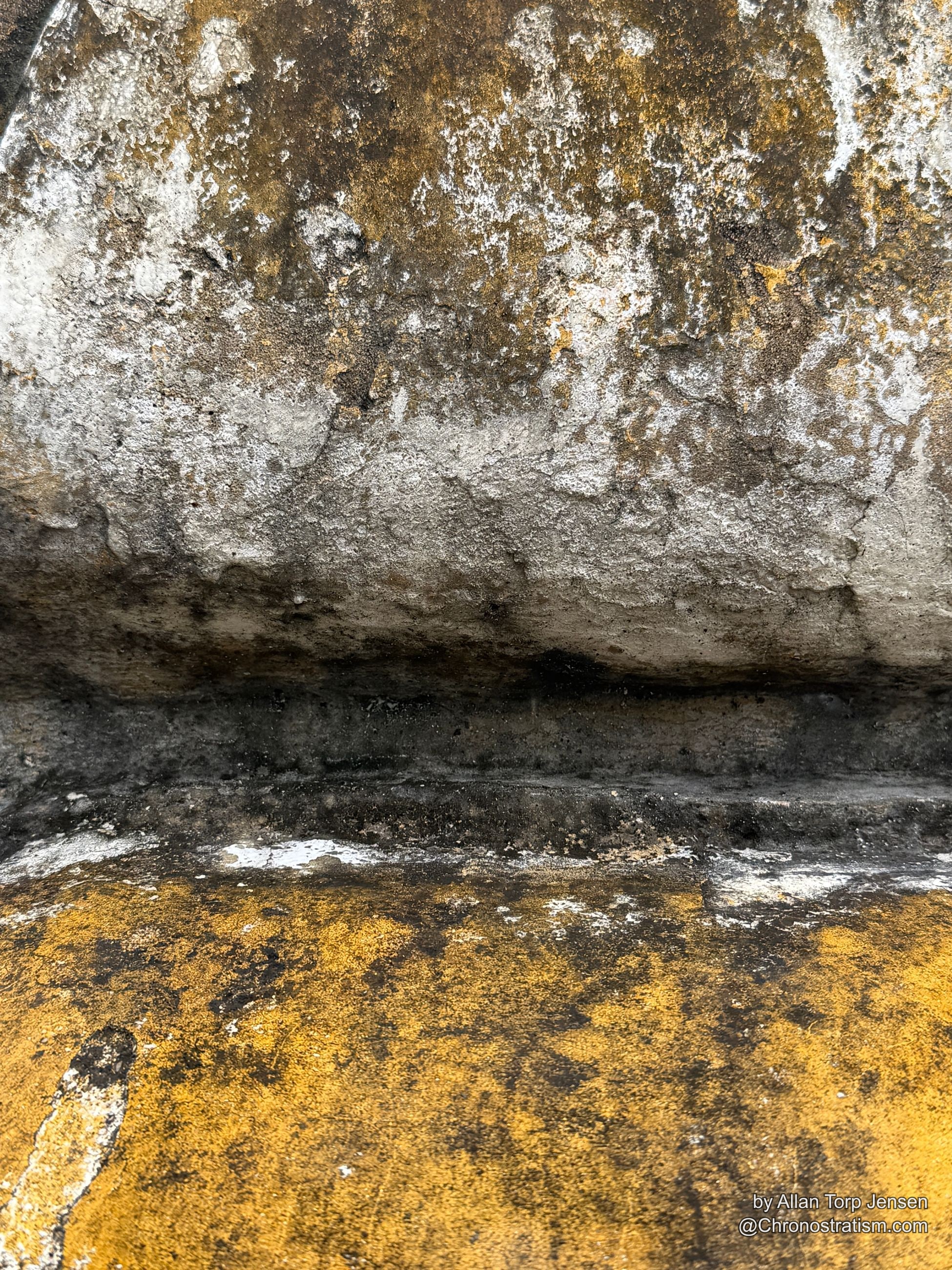 The gilded statue’s base meets plain grey render, soot patina fading into bare concrete.