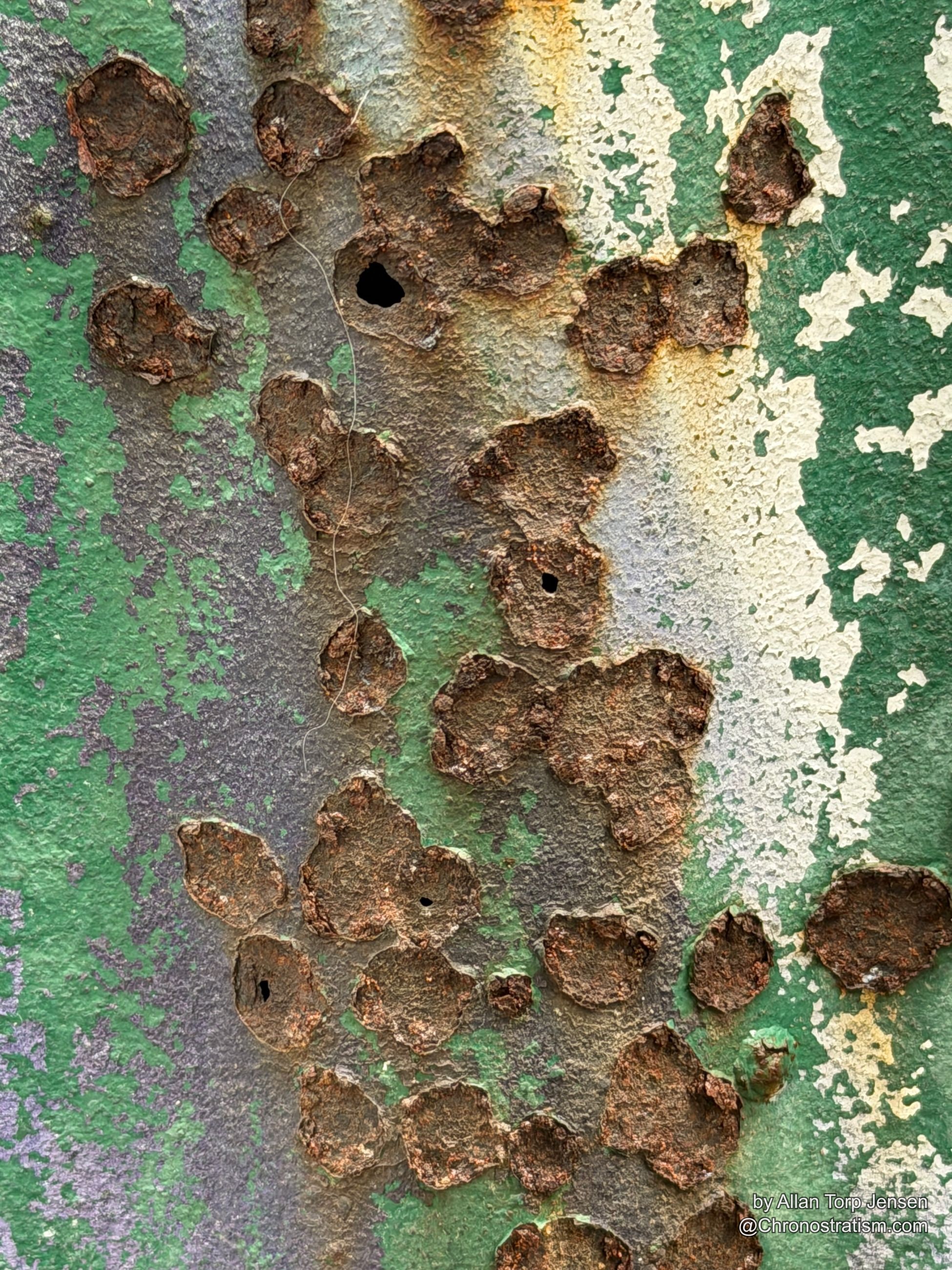 A jagged rust hole eats through pale blue-grey paint, metal curling back at the edges.