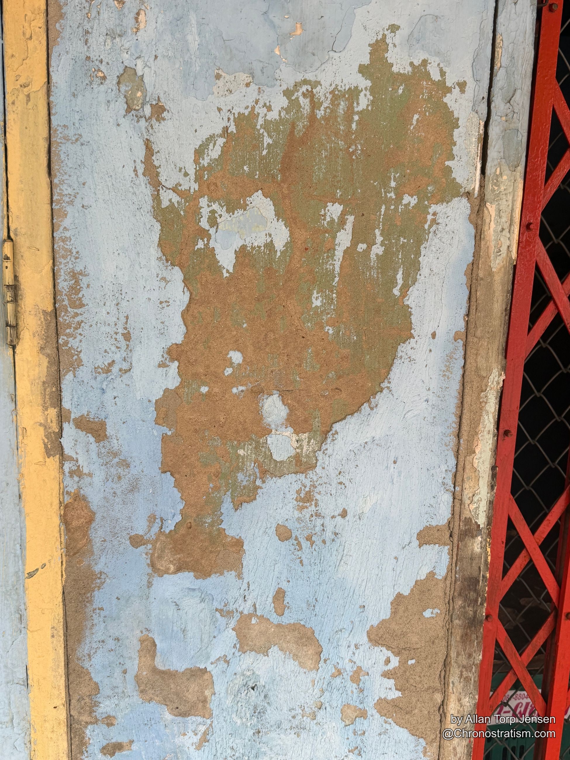 Layered blue-grey and tan paint peels back across weathered wood, a red mesh fragment caught at the edge.