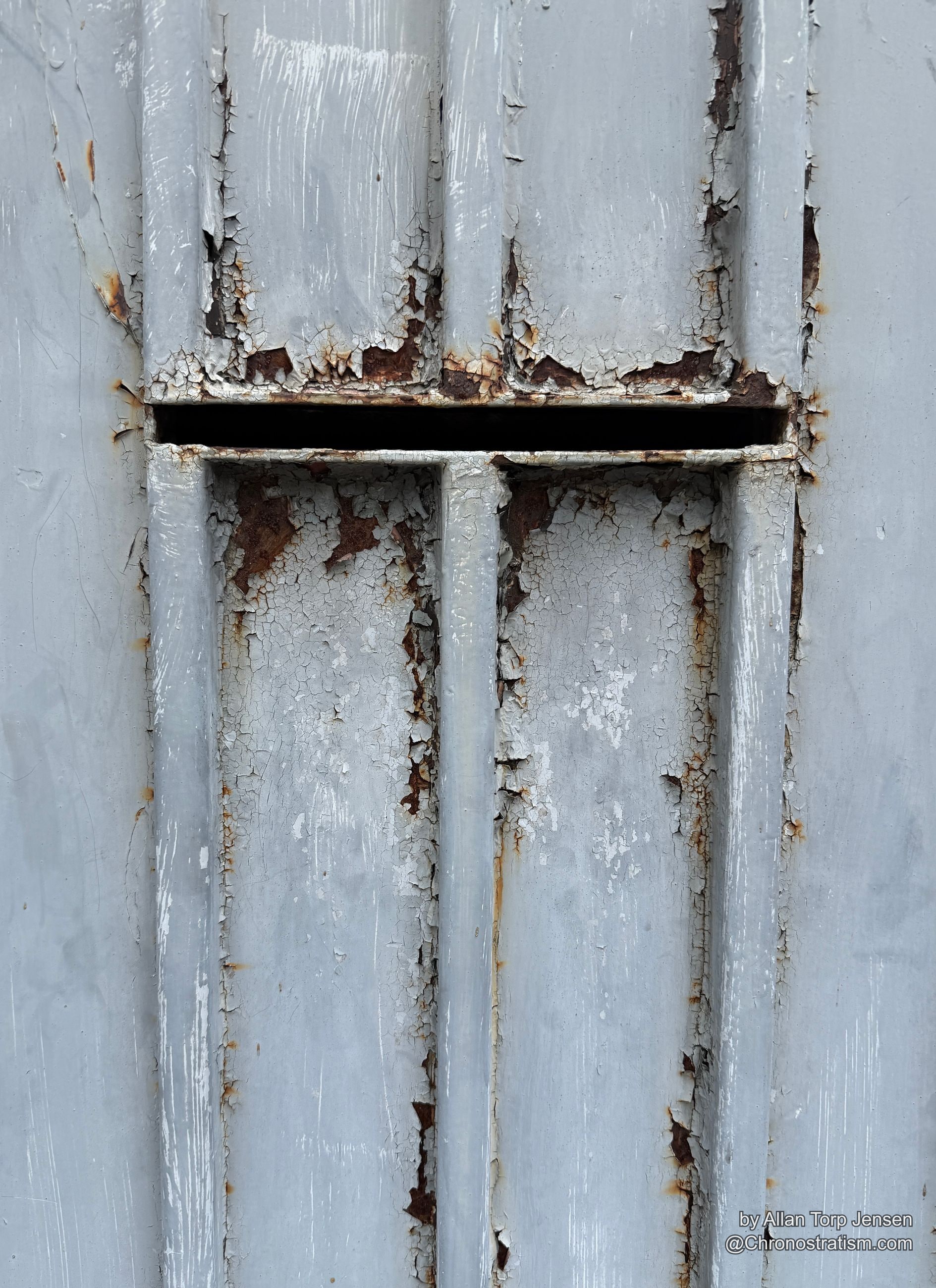 A pale grey door cracks and peels around a rusted mail slot, panel seams forming a cross.