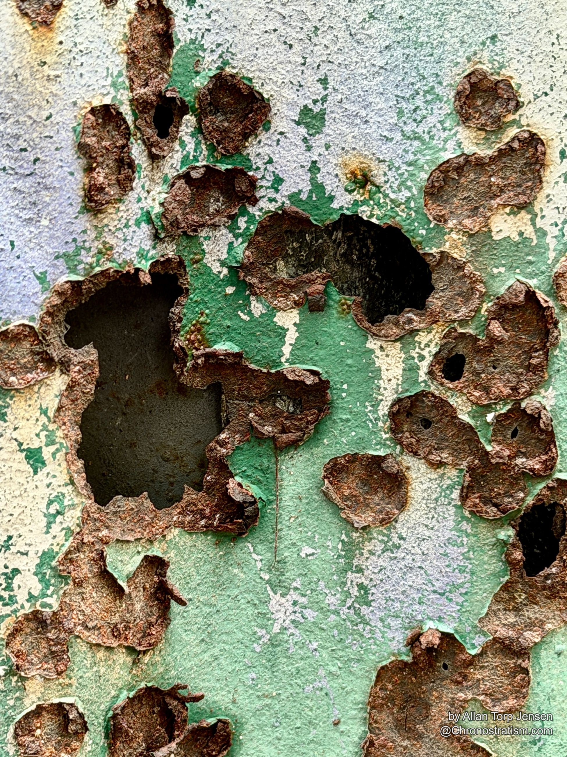 Purple and green paint peels from a heavily rusted metal panel, a bolt head rusting at its centre.