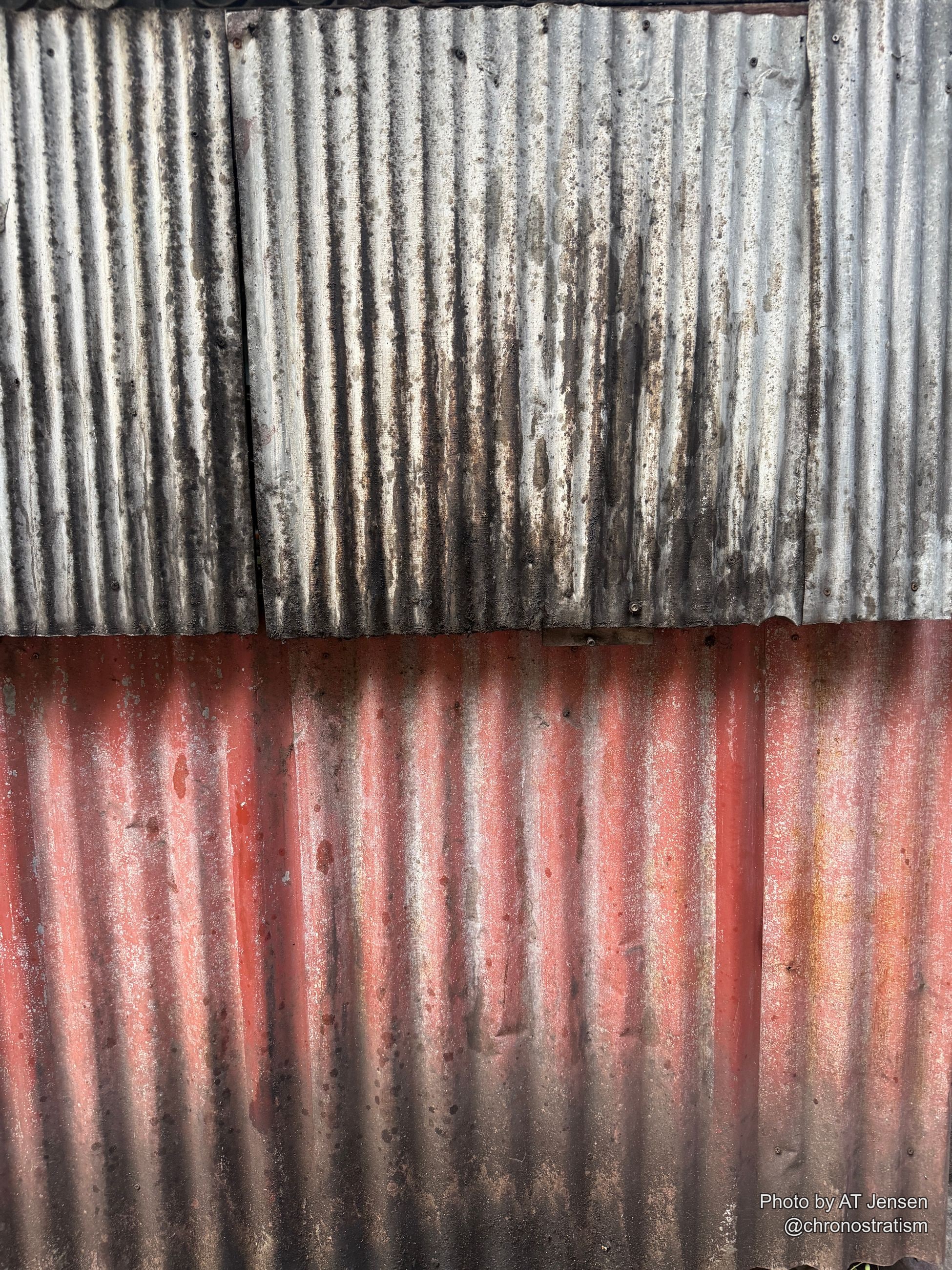 Rust bleeds through red paint on one half of a corrugated metal fence, soot streaking the other.