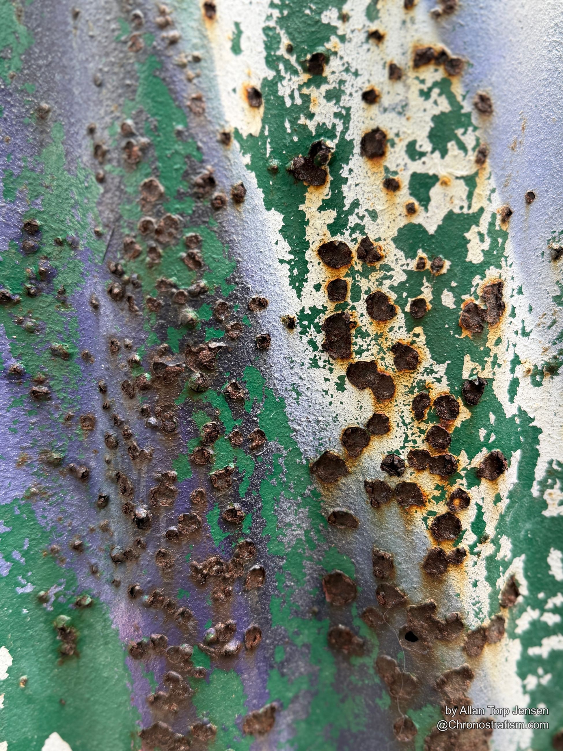 Rust eats clean through a spray-painted metal sheet, dozens of small perforations clustered together.
