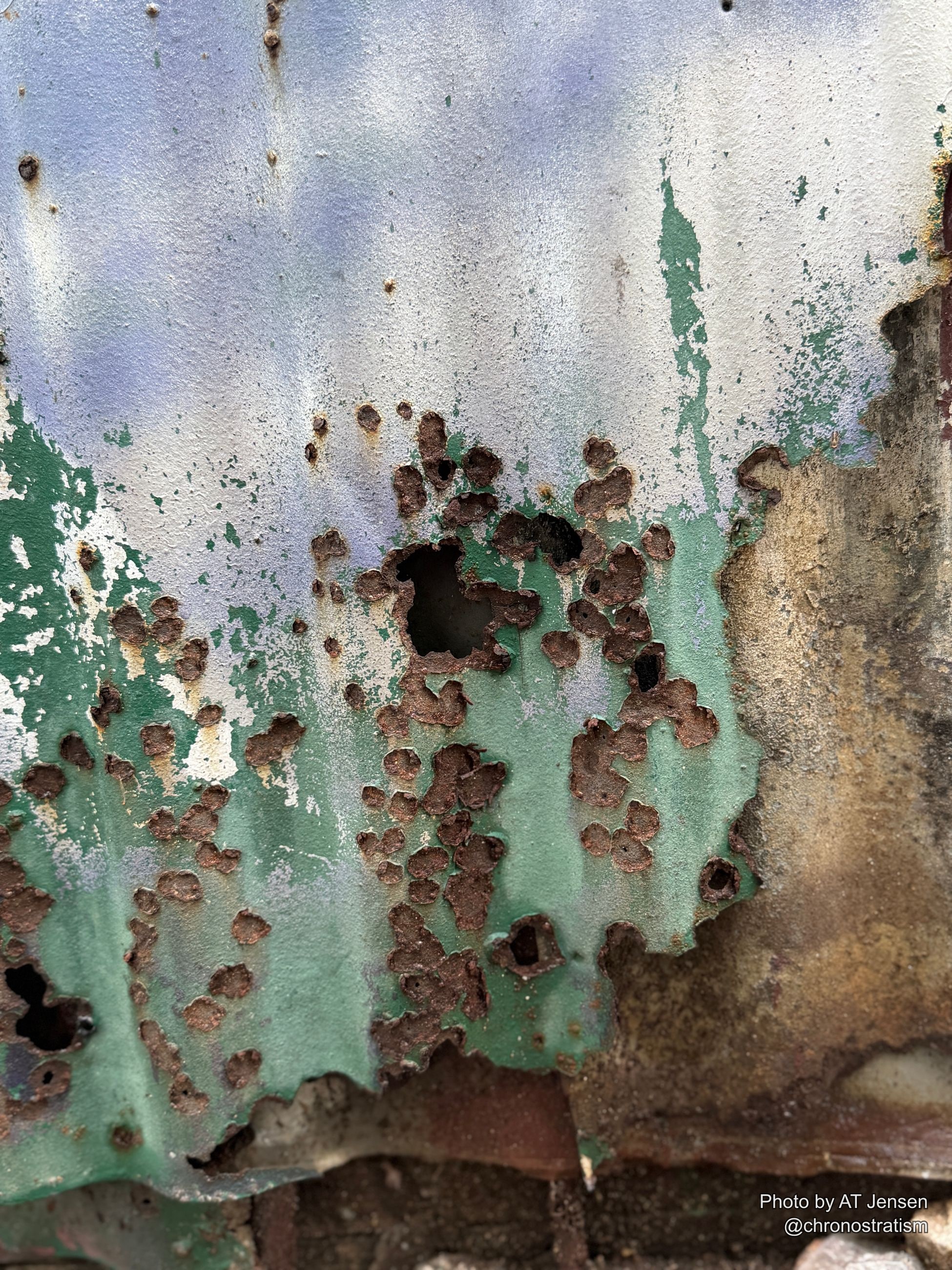 Rust holes cluster densely where green paint has failed on corrugated sheet metal.