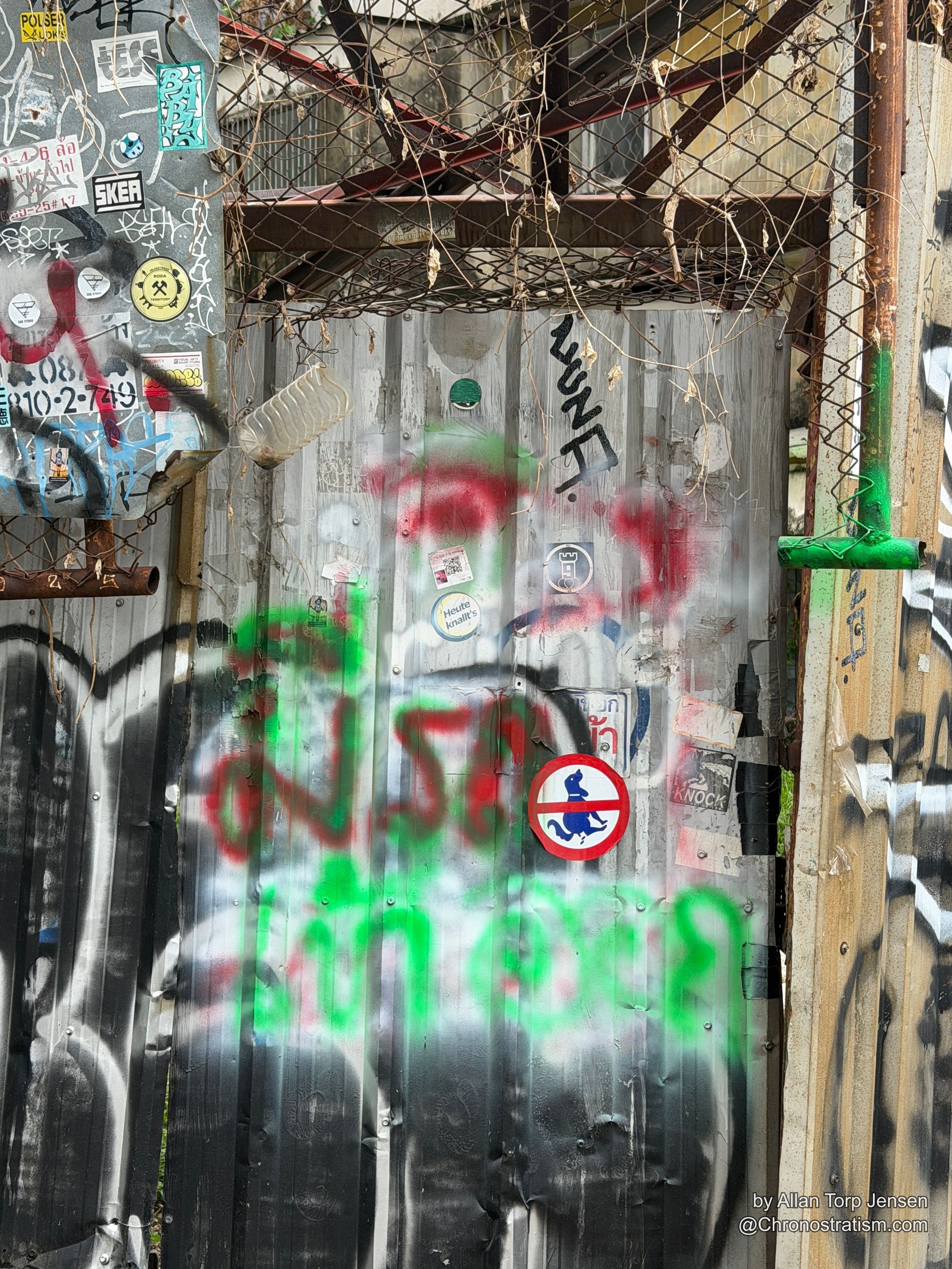 A rust-streaked corrugated wall layered with stickers, spray tags and a chain-link fence overlay.