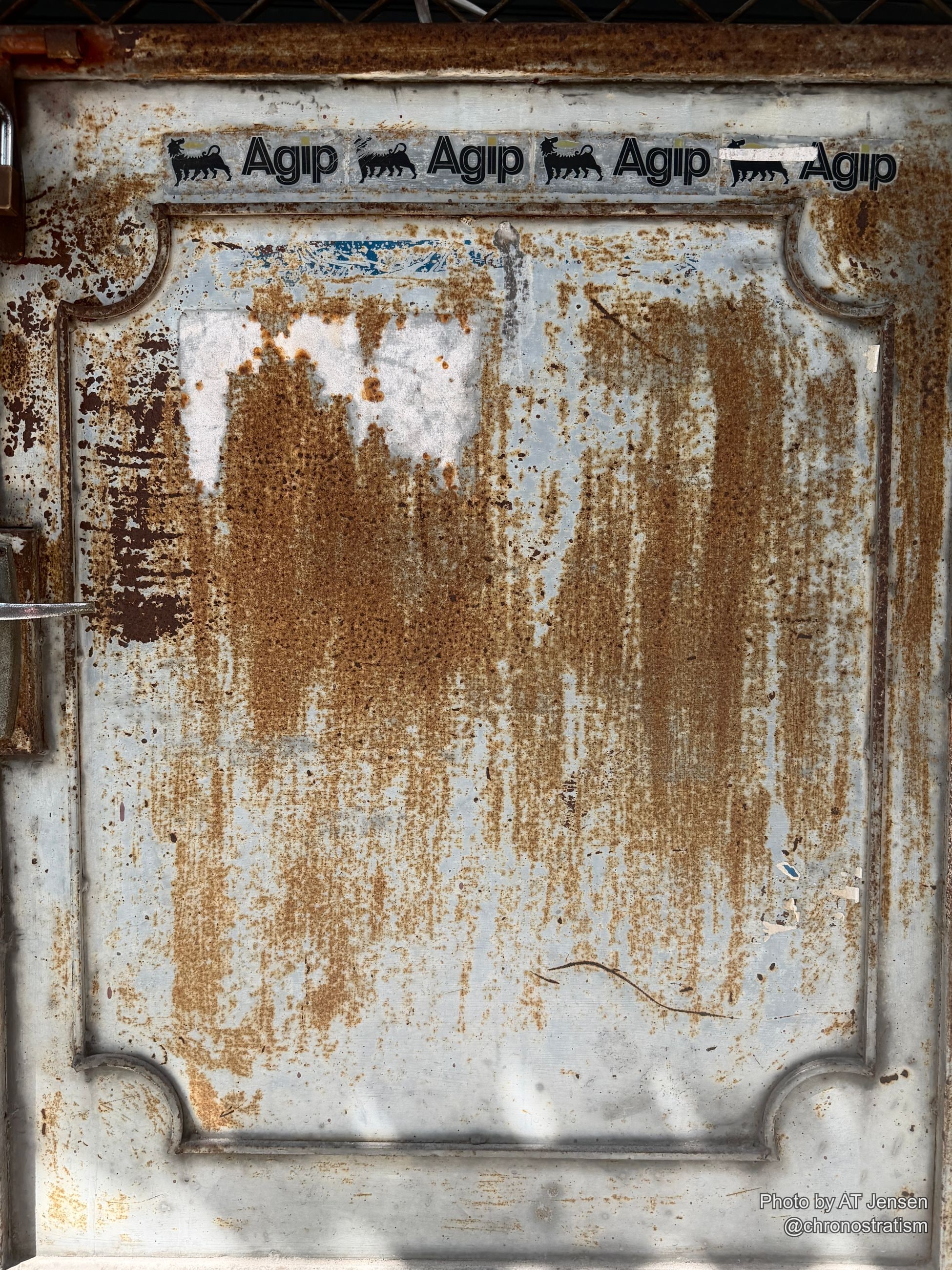 Rust streaks bleed down a metal door plastered with repeated Agip oil-brand stickers.