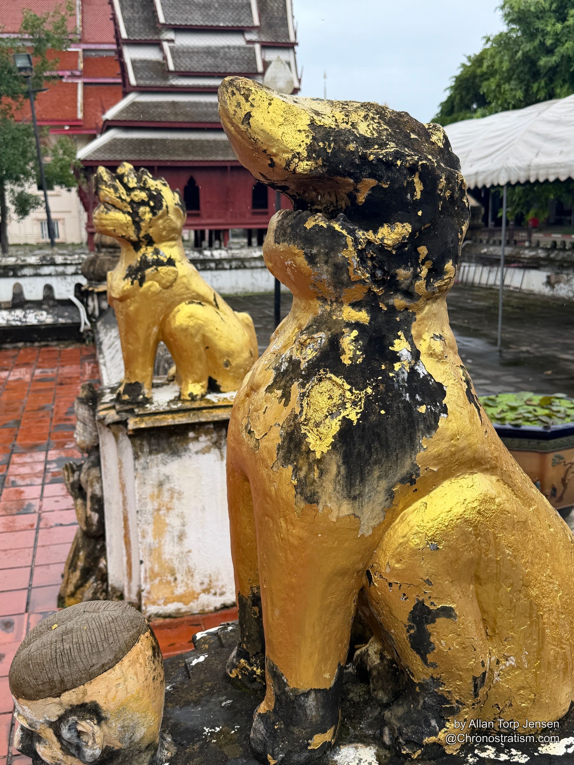 A soot-blackened gilded guardian statue sits before a Thai temple’s red rooftops, a twin figure visible behind.