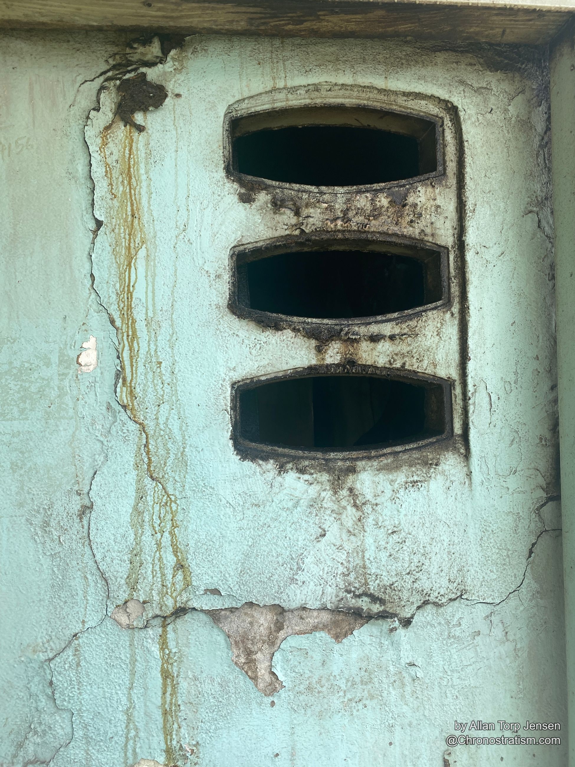 Soot-blackened ventilation slots cut into a cracked, teal-painted render wall.