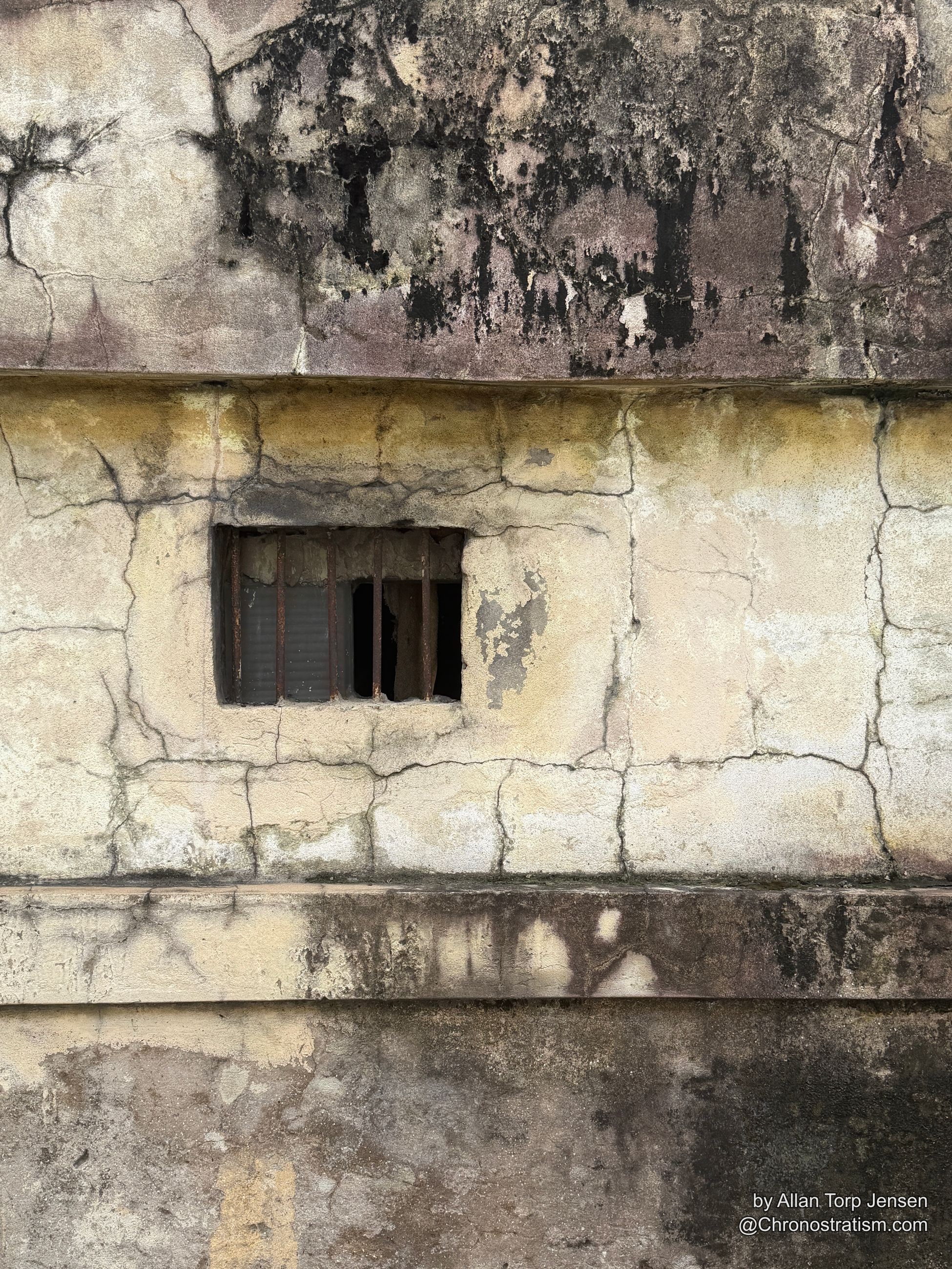 A weathered stucco pillar with a barred vent window, dark mould blotches spreading either side.
