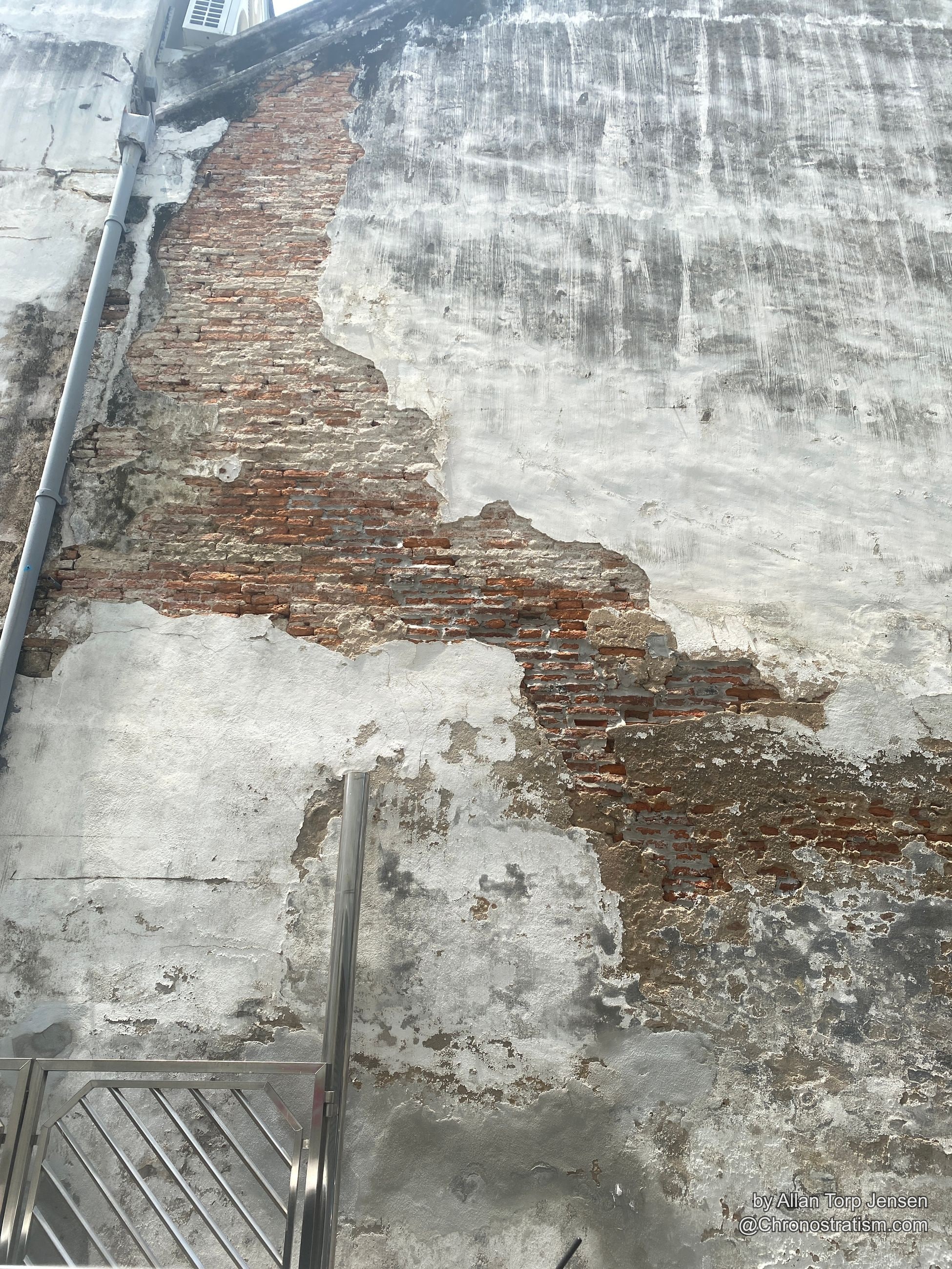 Weathered white render peeling back to raw brick, mottled staining across the wall.